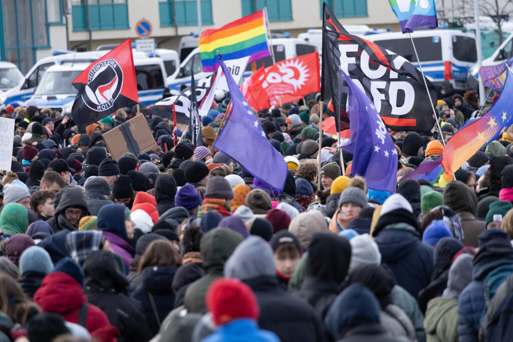 Viele demonstrierende stehen zusammen. Über ihren Köpfen wehen viele verschiedene Antifa, Volt und Pride Fahnen. Im Hintergrund parkende Polizeifahrzeuge