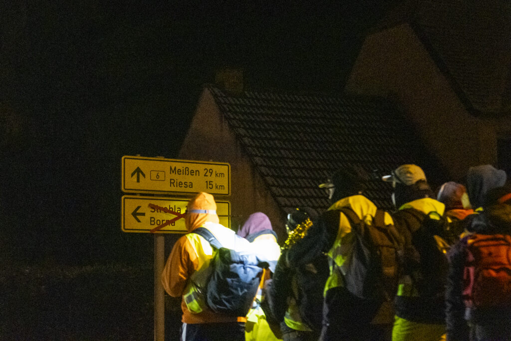 Ein paar Menschen stehen bei Dunkelheit vor einem Straßenschild der Bundesstraße B6. Das Schild weist auf Meißen in 29 Km und Riesa in 15 Km hin.