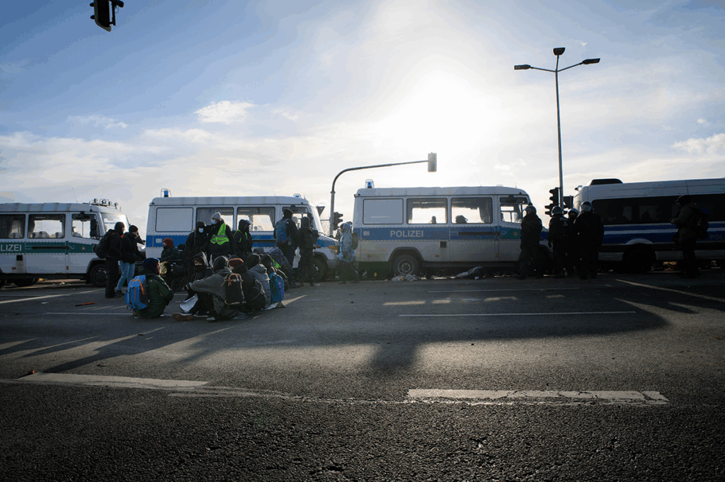 eine kleine Gruppe Menschen sitzt auf einer Straße vor einer Reihe Mannschaftswagen der Polizei. Vereinzelt stehen Polizeibedienstete herum. Die Sonne scheint.