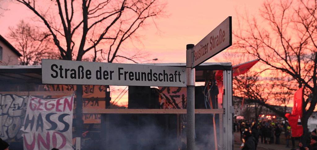 Im Hintergeund ein Lautsprecherwagen mit Bannern. Die Banner sind nicht vollständig lesbar. Im Vordergrund ist ein Straßenschild "Straße der Freundschaft"