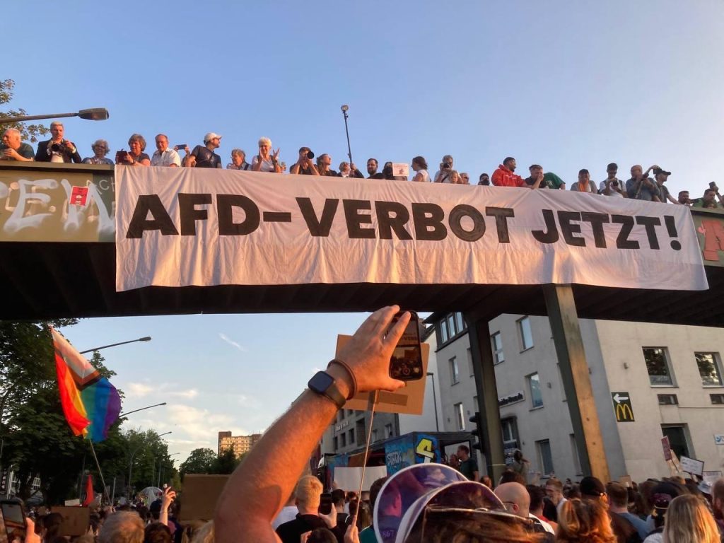 Eine mit Menschen gefüllte Brücke. Ein weißes Banner mit schwarzer "AFD-Verbot Jetzt!" Aufschrift hängt daran. Unter der Brücke eine demonstrierende Menschenmenge. Im Hintergrund Wohnhäuser.