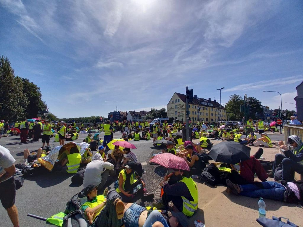 Viele Menschen mit Warnwesten sitzen bei Sonnenschein auf einer innerstädtischen Kreuzung. Im Hintergrund stehen vereinzelt Mannschaftswagen der Polizei.