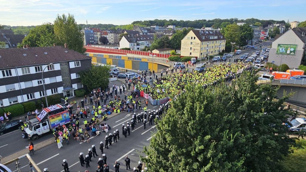 Luftaufnahme von einer Demo, die von Polizei umgeben ist. Vor der Demo sitzen einige Menschen auf dem Boden hinter einem Lautsprecherwagen. Der Lautsprecherwagen ist mit Bannern behängt. "FCK AFD" und "One Struggle One FIght, Reclaim, Resist, Revolt" ist auf diesen zu lesen.