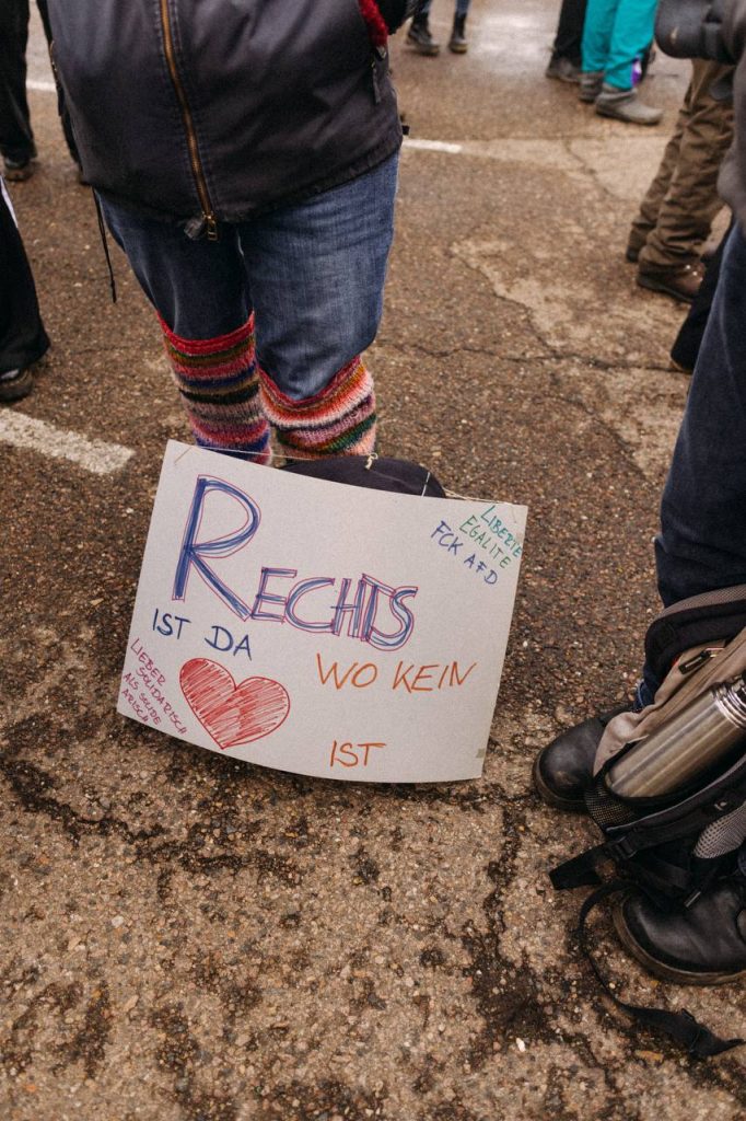 Ein gemaltes Schild lehnt an einem Rucksack, vor den Beinen einer Person. Auf dem Schild steht "Rechts ist da wo kein Herz ist. Liberte, Egalite, FCK AFD. Lieber Solidarisch als solide arisch".