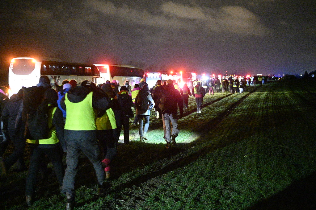 Viele Demonstrant*innen laufen bei Dunkelheit über ein Feld an stehenden Reisebussen vorbei. In Fahrtrichtung der Straße ist der Himmel durch Blaulicht erleuchtet