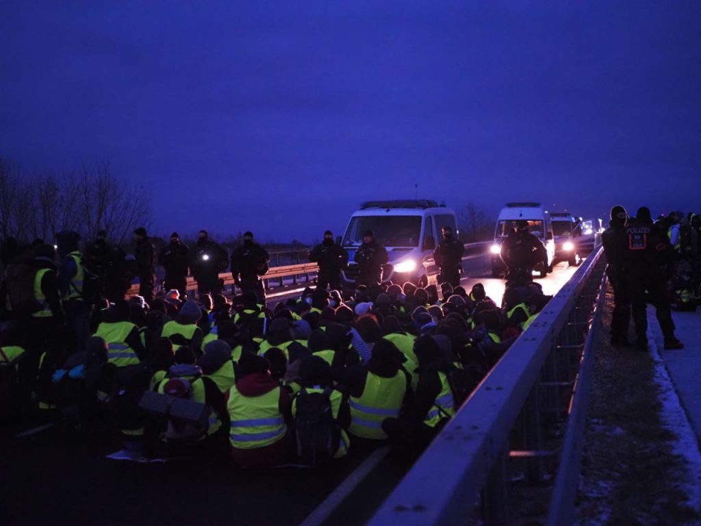 Eine Sitzblockade, die bei Dämmerung auf einer Straße sitzt. Die Polizei steht mit Fahrzeugen und Beamt*innen davor.